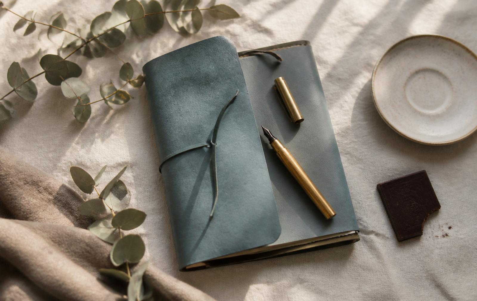 A teal notebook and an open journal with a fountain pen on warm ivory linen