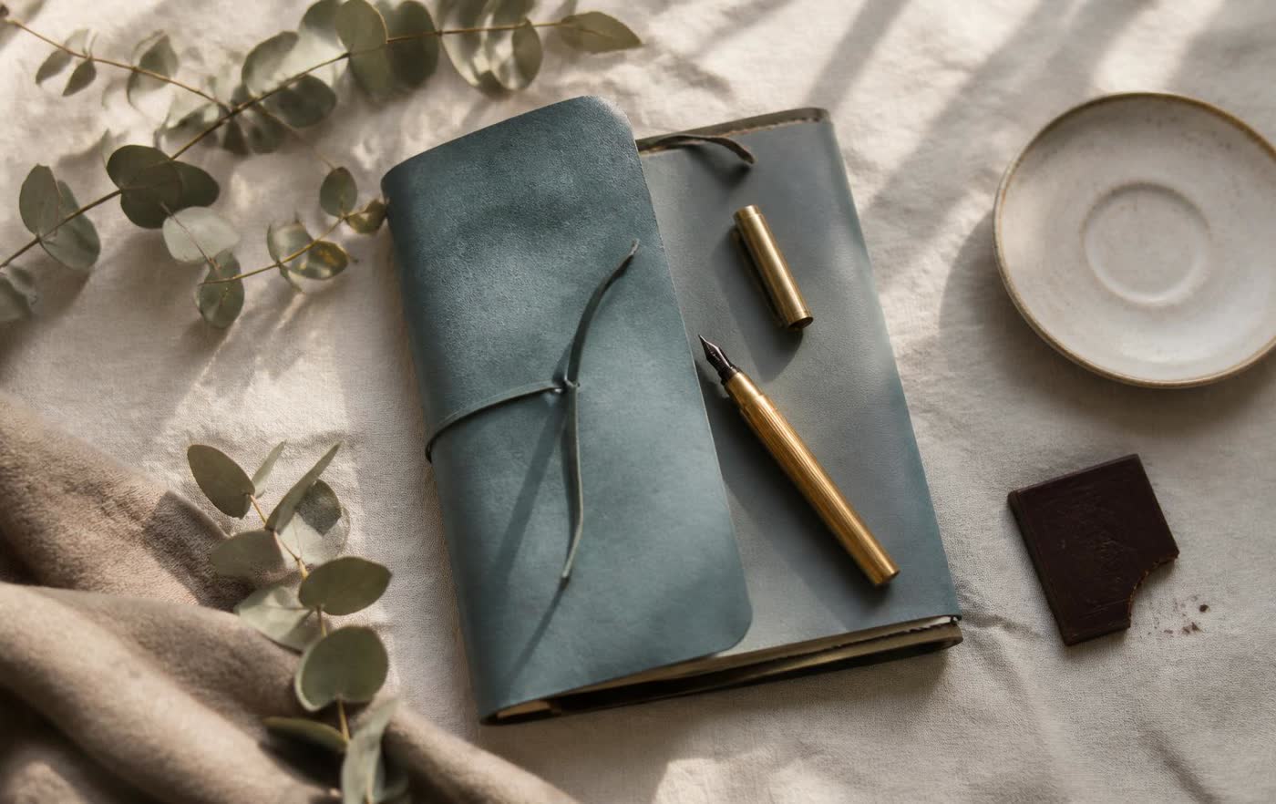 A teal notebook and an open journal with a fountain pen on warm ivory linen